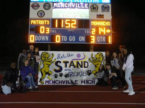 Menchville students and principal pose in front of the scoreboard before half-time.
